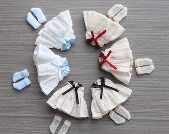 May include: A collection of six miniature doll dresses with eyelet lace fabric. Two white dresses feature light blue ribbons, two cream dresses have red ribbons, and two cream dresses have black collars with black ribbons. Includes matching socks.