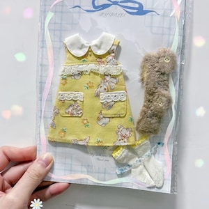 Pode incluir: Um vestido amarelo sem mangas com gola branca, acabamento em renda e estampa de coelhos, embalado com um cachecol de pele sintética marrom e meias brancas. A embalagem apresenta o texto PY DOLLS e Thank you.