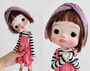 May include: A doll with short brown hair and a purple knit headband, wearing a black and white striped long-sleeve shirt under pink overalls with metal buckles. The doll has large dark eyes, rosy cheeks, and tan canvas sneakers.