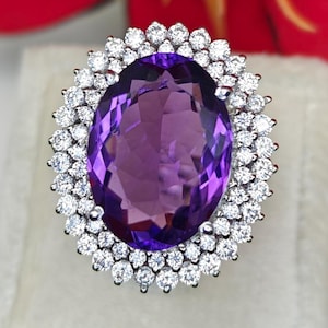 May include: An amethyst ring featuring a large, oval purple gemstone encircled by small, round, white gemstones. The ring is set on a white surface, with a red and orange flower in the background. The ring is a piece of jewellery.