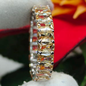 Puede incluir: Un anillo plateado con una banda continua de piedras preciosas rectangulares de color amarillo claro. El anillo está engastado sobre un fondo rojo y verde, con una tela blanca en primer plano. Las piedras preciosas están espaciadas uniformemente y reflejan la luz.