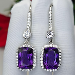 Puede incluir: Un par de pendientes colgantes de plata con grandes piedras rectangulares de color púrpura intenso, rodeadas de piedras claras más pequeñas. Los pendientes tienen un poste largo y delgado adornado con piedras claras adicionales y una piedra clara redonda grande en la parte superior.