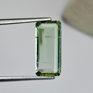 May include: A rectangular, light green gemstone with a faceted cut, held by a pair of silver tweezers. The gem has a subtle, translucent quality, with visible internal features. The edges of the gem are a darker shade of green.