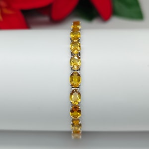May include: A silver bracelet featuring a series of oval-cut, golden-yellow gemstones. The gemstones are evenly spaced and set in a silver band. The bracelet is displayed against a white background with a hint of red and green floral elements.