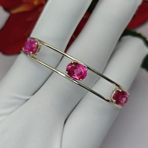 Op de afbeelding: Een zilverkleurige armband met een ontwerp van twee banden. Drie ovale, gefacetteerde felroze stenen zijn tussen de twee banden geplaatst, met één steen aan elk uiteinde en één in het midden, wat zorgt voor een gebalanceerde uitstraling.