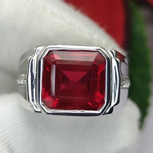 May include: A silver-toned ring featuring a large, square-cut, deep red gemstone set in the centre. The band has a polished metallic finish with small, clear accent stones embedded along the sides of the shank, framing the central red stone.