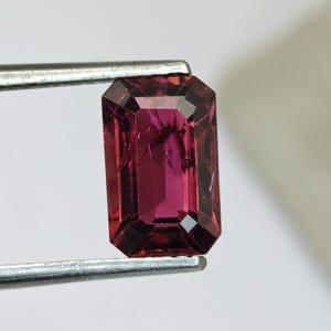 May include: A faceted, rectangular gemstone held by metal tweezers. The gem is a deep, rich magenta color with visible internal inclusions. The cut showcases multiple facets, reflecting light. The background is a plain white.