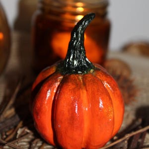 Op de afbeelding: Een kleine, oranje pompoenfiguur met een donkergroene en zwarte steel. De pompoen staat op een geweven oppervlak met twijgen en een vage achtergrond. Een herfstdecoratie.