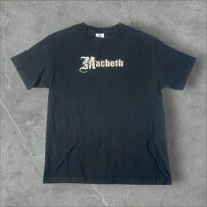 Puede incluir: Una camiseta negra de manga corta con la palabra Macbeth impresa en una fuente estilizada de inspiración gótica en el centro del pecho. El texto es principalmente blanco con detalles sutiles de desgaste y un pequeño acento rojo cerca de la b.