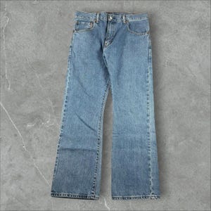 Könnte beinhalten: Eine mittelblaue Jeans aus Denim mit klassischem Fünf-Taschen-Design, Knopf- und Reißverschluss, Gürtelschlaufen am Bund und gerade geschnittenen Beinen mit sichtbaren, farblich abgestimmten Nähten, die das gesamte Kleidungsstück durchziehen.