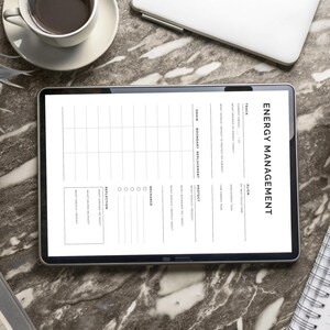 May include: A digital tablet displaying an "Energy Management" worksheet, with sections for tracking, reflection, and recharge. The tablet is surrounded by a coffee cup, a laptop, a notebook, and potted plants, all on a marble surface.