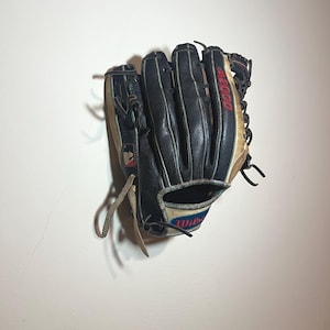 Puede incluir: Un guante de béisbol negro y marrón con detalles rojos y blancos. El guante presenta una etiqueta Wilson A2000 y un logotipo de la Major League Baseball. El guante está hecho de cuero y tiene un aspecto desgastado.