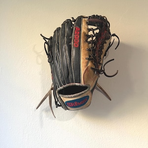 May include: A black and tan Wilson A2000 baseball glove. The glove has red stitching and lettering, with the Wilson logo in blue and red. The glove is hanging on a white wall.