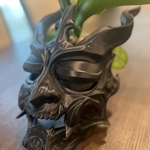 May include: A black, intricately detailed Oni mask, a traditional Japanese demon mask, with sharp teeth and horns. The mask is made of a matte material and is partially obscuring green plant leaves.