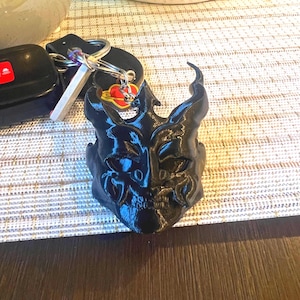 Może przedstawiać: Czarny brelok do kluczy z motywem stylizowanej maski. Maska ma skomplikowane detale i jest przymocowana do kółka na klucze z kluczami i pilotem do samochodu. Brelok leży na jasnej, wzorzystej powierzchni.