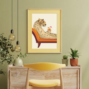 May include: A framed art print featuring a relaxed leopard lounging on an orange chaise lounge, holding a cocktail with a pink umbrella. The leopard is tan with black spots. The print has a yellow border.