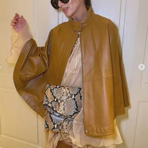 以下が含まれることがあります： ハイカラーとフロントジッパーが付いたタンレザーケープジャケット。シルバーのハードウェアが付いたスネークスキンプリントのクラッチが、透明で明るい色のドレスの上に着用されています。モデルは濃い色のサングラスをかけています。