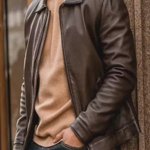 Op de afbeelding: Een donkerbruine leren jas met een klassieke kraag en een ritssluiting over de gehele lengte aan de voorzijde. De jas wordt gedragen over een beige trui met ronde hals en een donkere spijkerbroek. Het leer heeft een gladde afwerking met zichtbare naden.