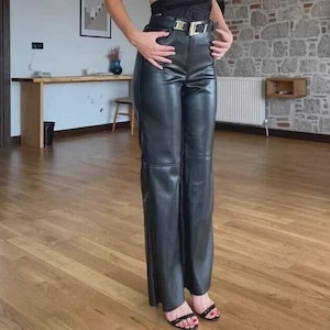 Peut inclure: Pantalon noir taille haute avec une finition lisse, une coupe large et une couture horizontale au niveau des cuisses. Le pantalon est maintenu à la taille par une ceinture noire dotée d'une boucle en métal argenté bien visible sur le devant.