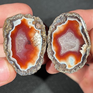 May include: Two halves of a polished agate stone, showcasing a vibrant red and white interior with a crystalline center. The outer edges are a mix of brown and gray, creating a natural, earthy aesthetic. The stones are held in a person's hands.