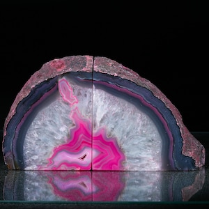 Op de afbeelding: Twee agaat boekensteunen, elk een halfronde vorm. De buitenrand is een ruw, donkerroze. De binnenkant heeft lagen van donkerblauw, wit en levendig roze. De boekensteunen worden op een reflecterend oppervlak weergegeven.