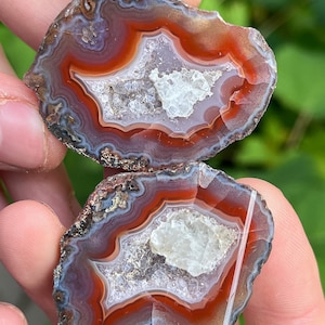 May include: Two halves of a polished agate stone, showcasing concentric bands of red, white, and gray hues. The center of each stone features a crystalline cavity. The natural stone is held in a person's hand.