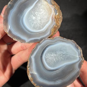 May include: Two halves of a polished agate stone, showcasing concentric bands of gray, white, and translucent hues. The center of each piece features a crystalline formation. The outer edges have a rough, brown texture.
