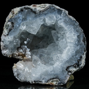 May include: A geode with a hollow center filled with light blue and clear crystal formations. The outer shell is a mix of gray, white, and black, creating a natural, textured appearance. The geode is set against a black background.