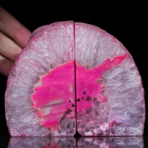 May include: Two agate bookends, each cut in a semi-circular shape. The bookends have a bright pink centre with white and translucent outer edges. The natural stone has a polished surface and is set against a black background.
