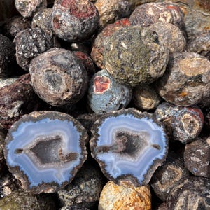 Könnte beinhalten: Eine Ansammlung runder, rau strukturierter Geodenknollen in Grau-, Braun- und Rosttönen, wobei zwei Hälften einer gespaltenen Geode im Vordergrund konzentrische hellblaue und weiße Achatbänder um einen zentralen Kristallhohlraum zeigen.