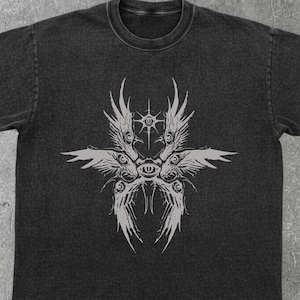 Puede incluir: Camiseta gris oscuro con acabado desgastado. La camiseta presenta un gráfico simétrico y estilizado en gris claro, que se asemeja a una criatura celestial o mítica con alas y ojos. El diseño está centrado en la parte delantera de la camiseta.