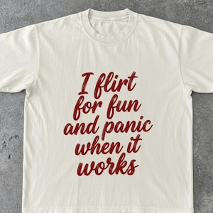 May include: Cream-colored t-shirt with the phrase "I flirt for fun and panic when it works" in a burgundy script. The shirt has a crew neck and short sleeves, suitable for casual wear.