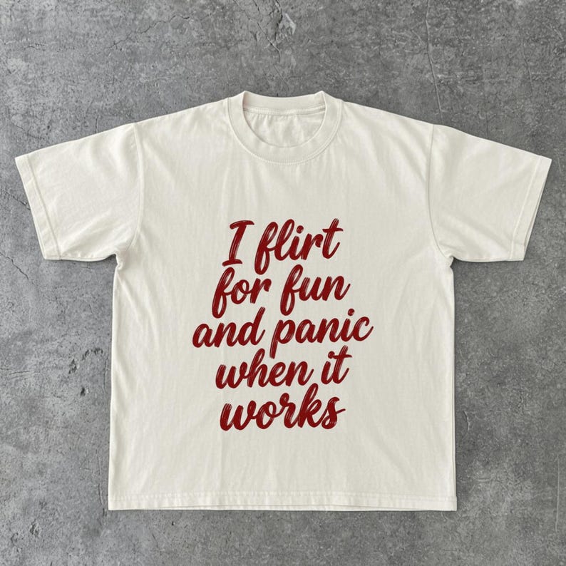 May include: Cream-colored t-shirt with the phrase "I flirt for fun and panic when it works" in a burgundy script. The shirt has a crew neck and short sleeves, suitable for casual wear.