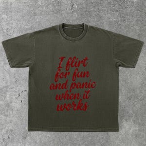 May include: Olive green t-shirt with the red text "I flirt for fun and panic when it works." The shirt has short sleeves and a crew neck. The fabric appears to be a soft cotton blend.