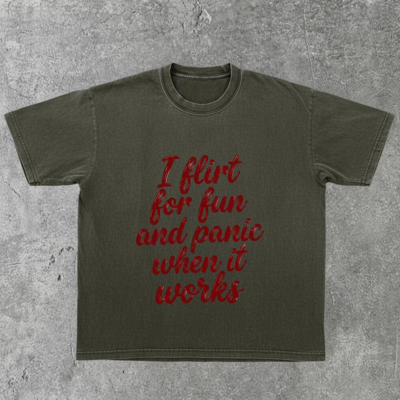 May include: Olive green t-shirt with the red text "I flirt for fun and panic when it works." The shirt has short sleeves and a crew neck. The fabric appears to be a soft cotton blend.
