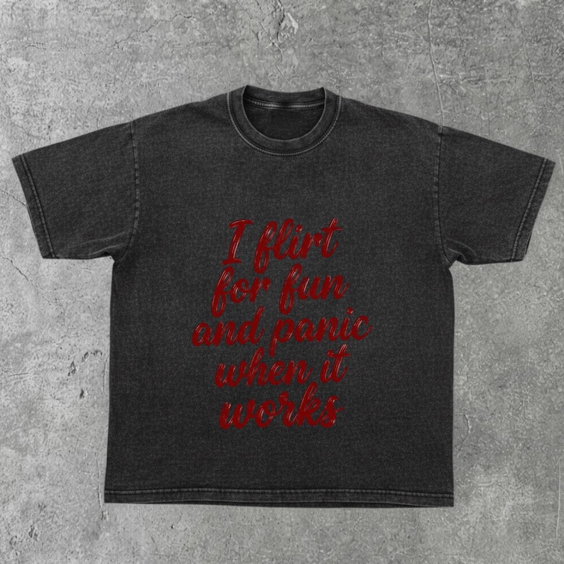 May include: Dark gray t-shirt with the text "I flirt for fun and panic when it works" in red script. The shirt has a vintage, washed appearance and a crew neck.