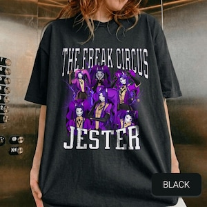 Può includere: T-shirt nera con la scritta "THE FREAK CIRCUS" e "JESTER" a blocchi bianchi. La grafica presenta un gruppo di giullari dai capelli viola in abiti viola e oro. La parola "BLACK" è in un rettangolo nero in basso.