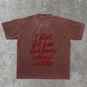 May include: A brown t-shirt with the text "I flirt for fun and panic when it works" in red script. The shirt is a solid color and has a crew neck.