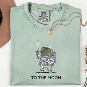 Puede incluir: Una taza de cerámica blanca con asa negra y el texto "But First, Coffee" en negrita negra. La taza está sobre un fondo borroso. La taza mide aproximadamente 10 cm de altura.