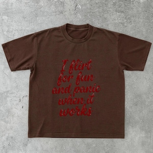 May include: A brown t-shirt with the red text "I flirt for fun and panic when it works." The shirt has short sleeves and a crew neck. The text is in a cursive font.