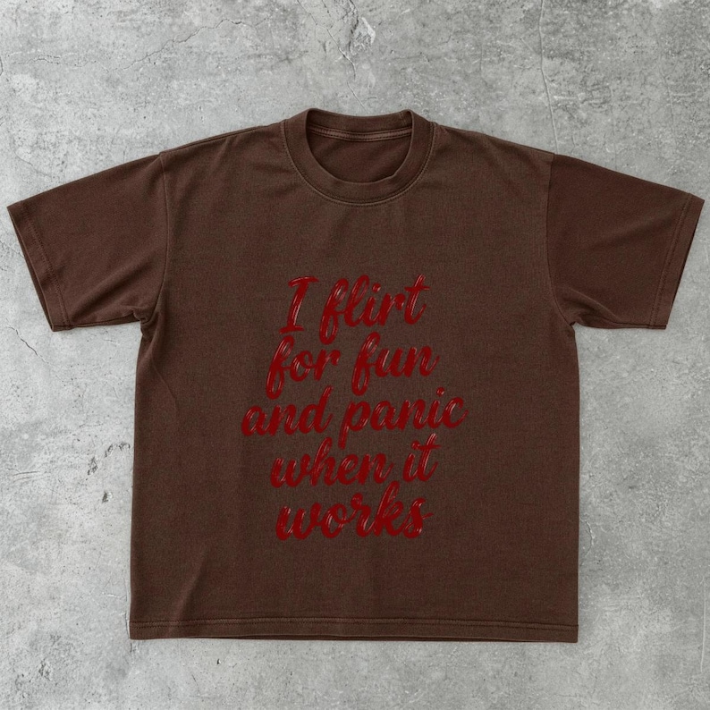May include: A brown t-shirt with the red text "I flirt for fun and panic when it works." The shirt has short sleeves and a crew neck. The text is in a cursive font.