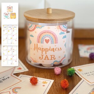Op de afbeelding: Een glazen pot met een houten deksel, met het opschrift "Happiness JAR" met een regenboogontwerp. De pot bevat opgerolde briefjes en is omgeven door kleurrijke pompons en kleine kaartjes. Het thema is positiviteit en welzijn.