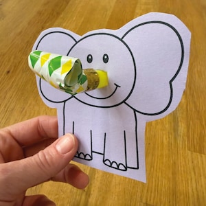 May include: A light purple elephant-shaped party favor with black outlines and a smiling face. A rolled-up party blower with yellow, green, and white diamond patterns is inserted into the elephant's trunk. The elephant is held by a hand.