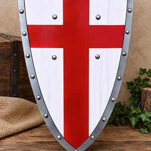 May include: A heater-style shield featuring a white surface with a bold red cross centered on the front. The shield is framed by a dark metallic border secured with evenly spaced silver rivets along the entire perimeter of the shield edge.