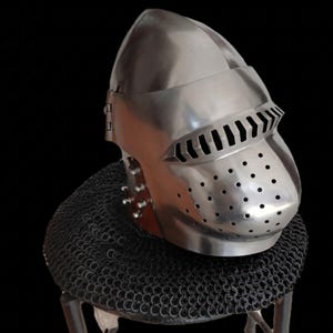 May include: A polished steel helmet with a visor and ventilation holes, displayed on a chainmail coif. The helmet features a rounded top and a hinged visor with horizontal slits. The chainmail is a dark grey colour.