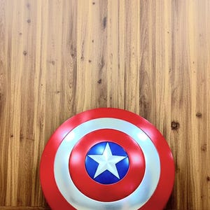 May include: A red, white, and blue shield with a star in the center. The shield is round with a silver ring around the center star. The background is a wooden wall and floor.
