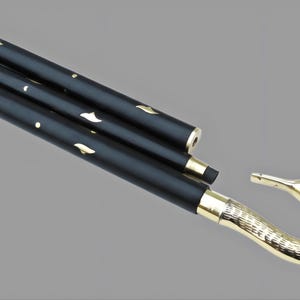 May include: A black folding cane with gold accents. The cane features a swan-shaped handle and decorative gold leaf-shaped cutouts. The handle and the ends of the cane are gold. The cane is folded and angled across a gray background.