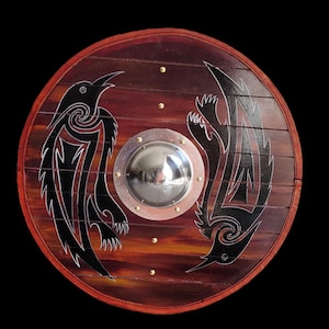 Puede incluir: Escudo redondo de madera con acabado marrón oscuro y borde rojo. El escudo presenta dos diseños estilizados de cuervos negros con contornos blancos. Un brocal de metal plateado está en el centro, y remaches de latón están colocados alrededor.