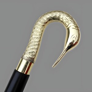 May include: A gold-colored walking cane with a bird-shaped handle. The handle has a textured pattern and a bird's head design. The shaft is black with a gold band near the handle. The cane is likely made of metal.