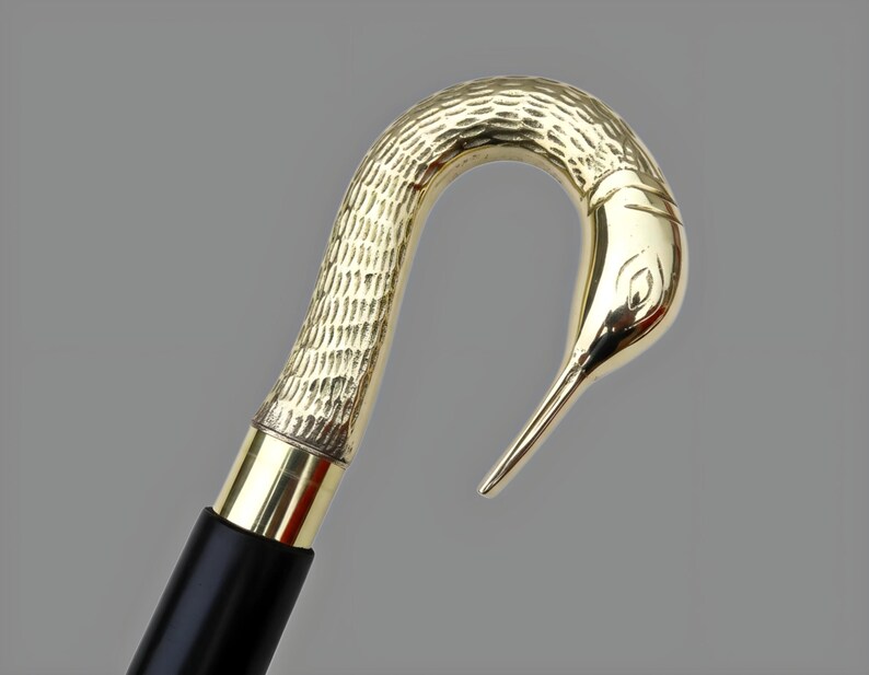 May include: A gold-colored walking cane with a bird-shaped handle. The handle has a textured pattern and a bird's head design. The shaft is black with a gold band near the handle. The cane is likely made of metal.
